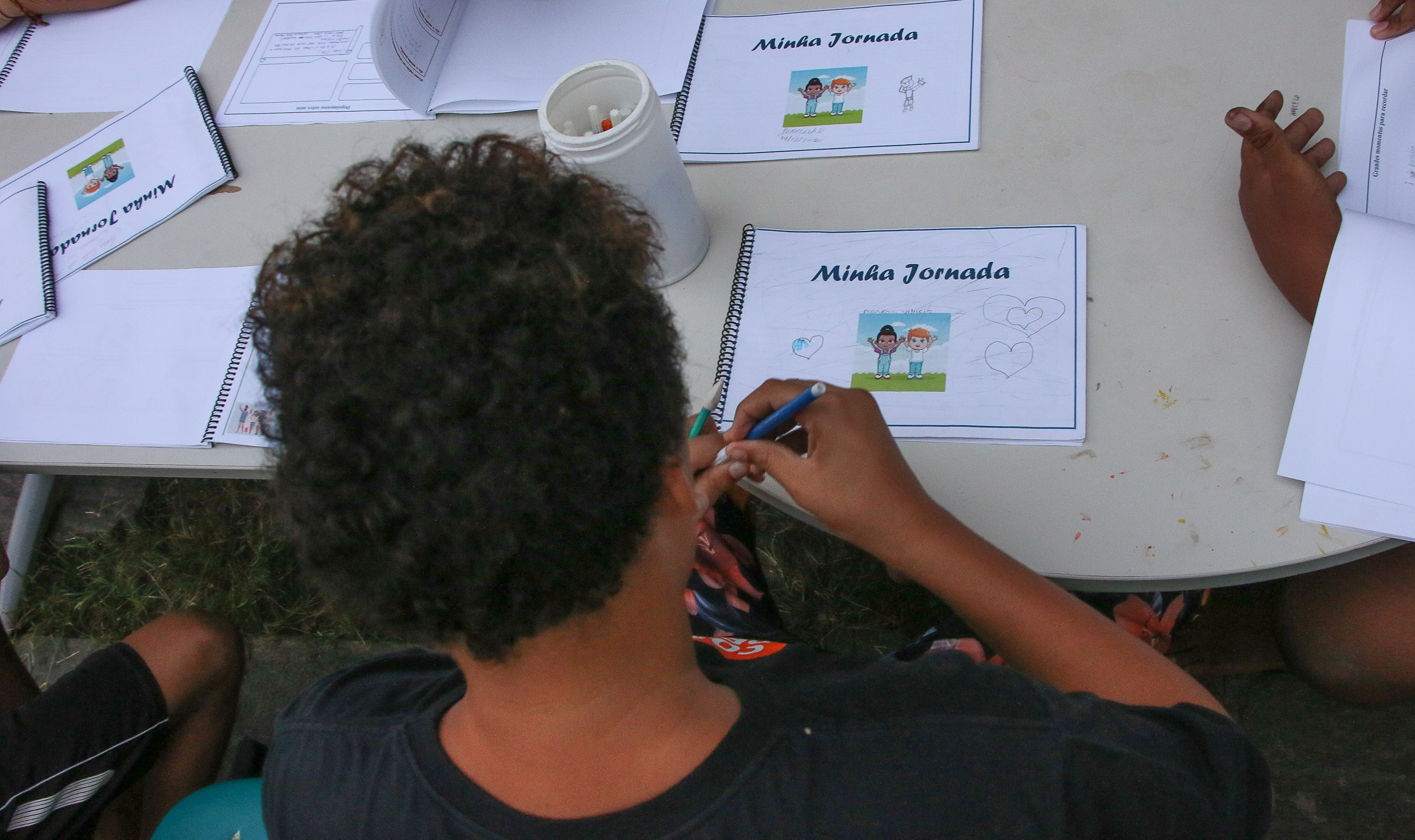 Na foto, um menino de costas e sem mostrar o rosto desenha em um caderno